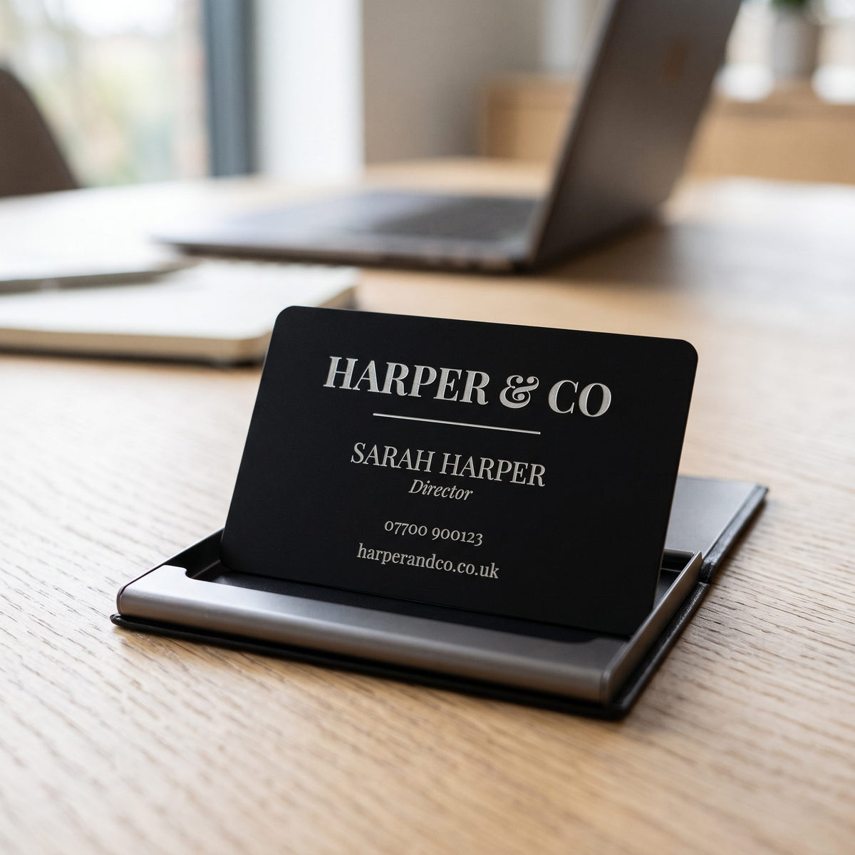 Black metal business card engraved Harper & Co in a card holder on a desk