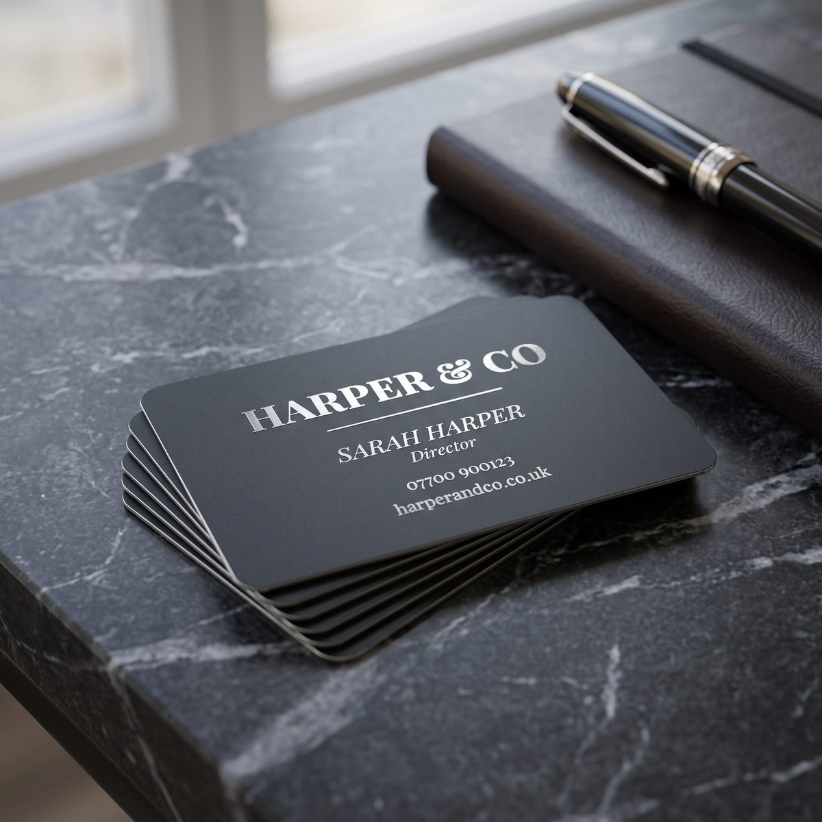 A stack of engraved black metal business cards on marble with a pen