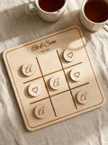 Personalised noughts & crosses set engraved with Ali & Sara, mid-game on a linen tablecloth with tea